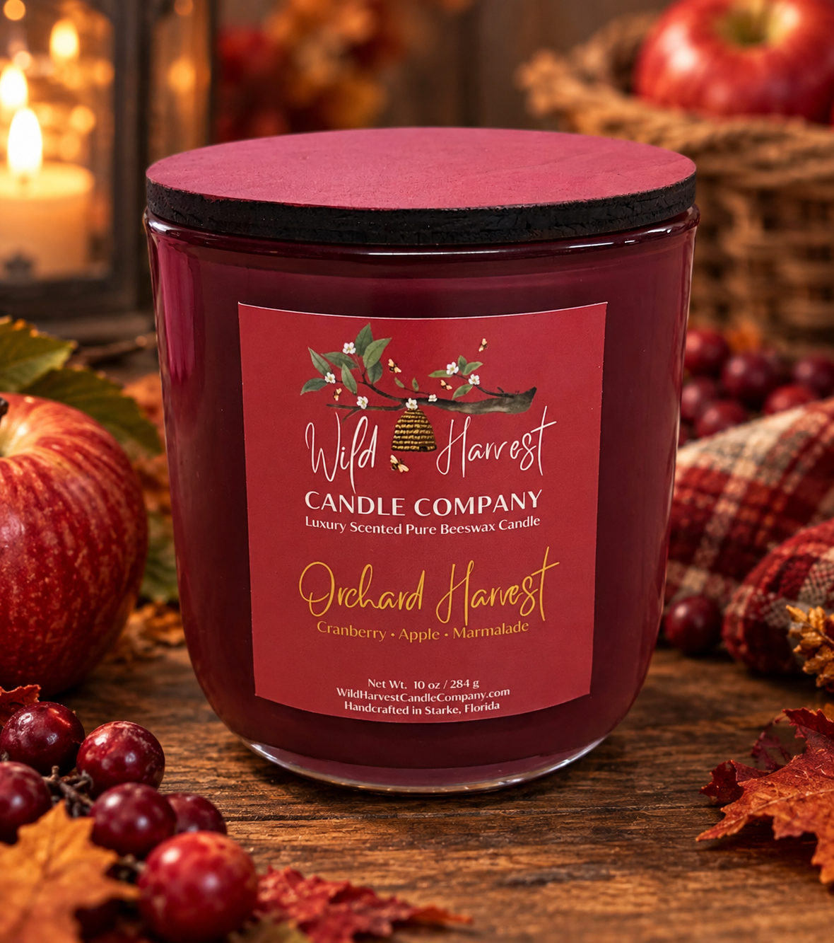 A jar candle from Wild Harvest Candle Company labeled 'Orchid Harvest' sits on a rustic wooden surface surrounded by fall-themed props including red apples, cranberries, leaves, and a lit candle in the background.