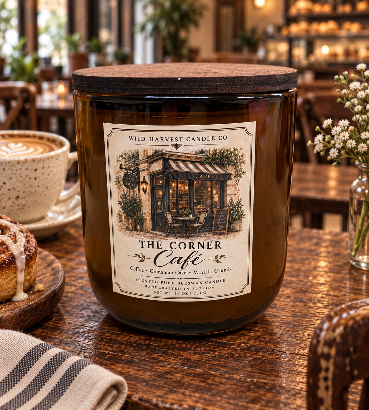 A large amber glass jar candle with a dark wood lid sits on a wooden table in a cozy cafe setting. The front label reads 'The Corner Cafe' and 'Wild Harvest Candle Co.