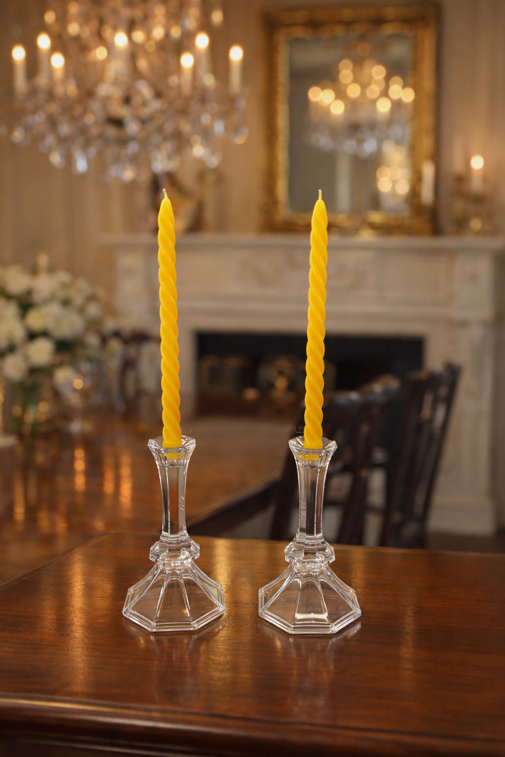 Two yellow candles in crystal candlesticks on a wooden table with a chandelier and mirror in the background.