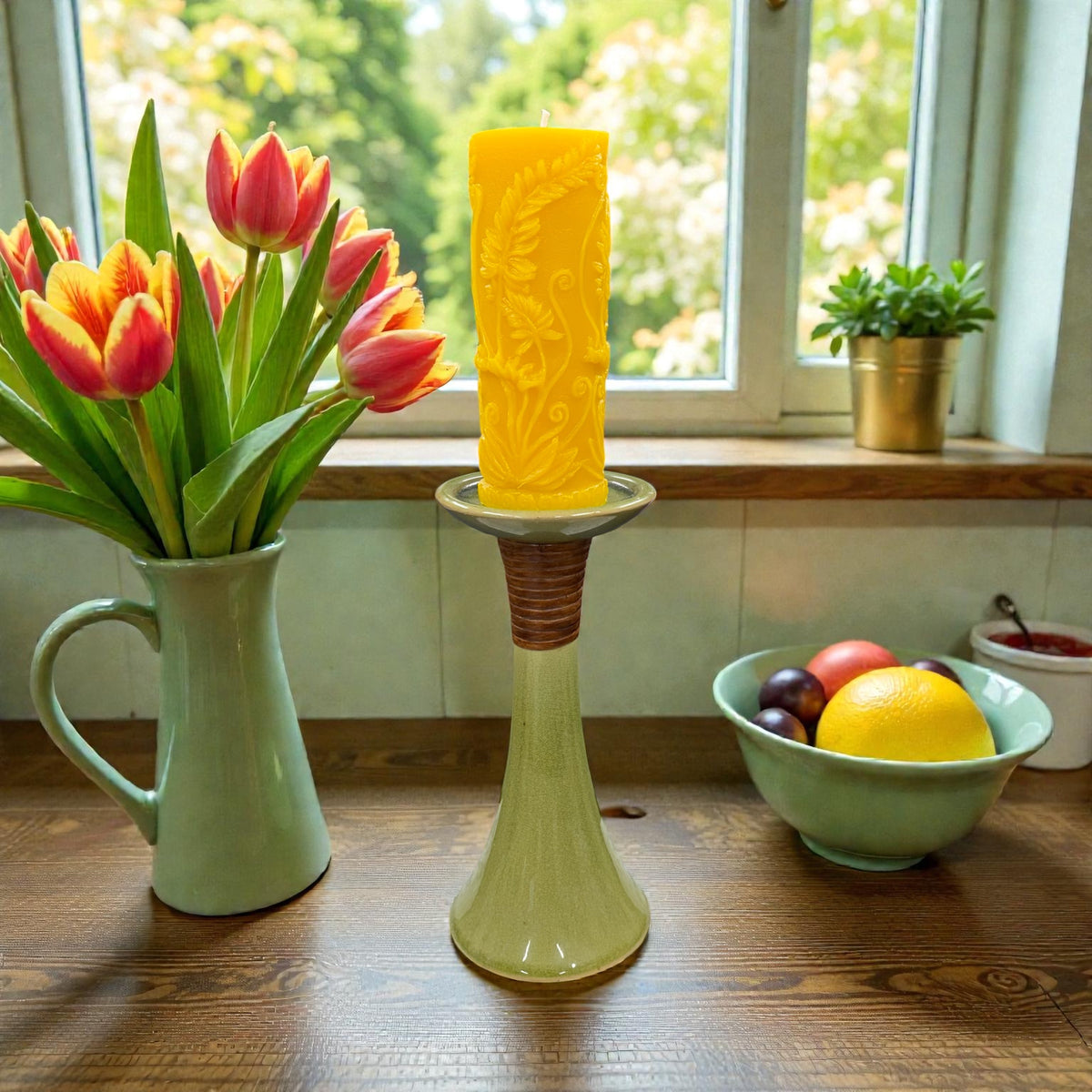 Yellow textured candle on a decorative stand with a vase of red tulips and a bowl of fruit in the background.