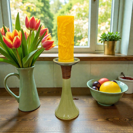Yellow textured candle on a decorative stand with a vase of red tulips and a bowl of fruit in the background.