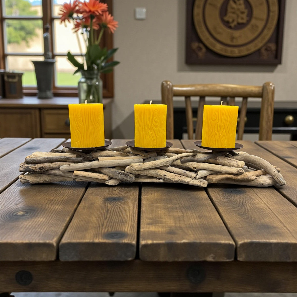 Three yellow candles on a wooden table with driftwood, in a room with a window and decorative items.